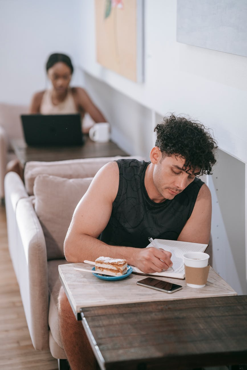 Person journaling about food with a relaxed expression at a sunny kitchen table with fresh fruit and coffee