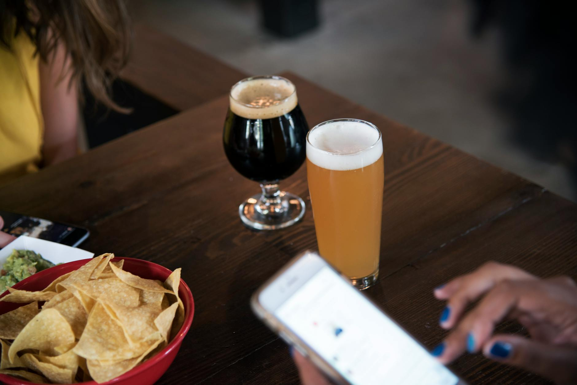 Person holding a schooner glass beside a smartphone with a calorie tracking app showing the kilojoule entry for the beer, on a wooden pub table with a small pretzel bowl
