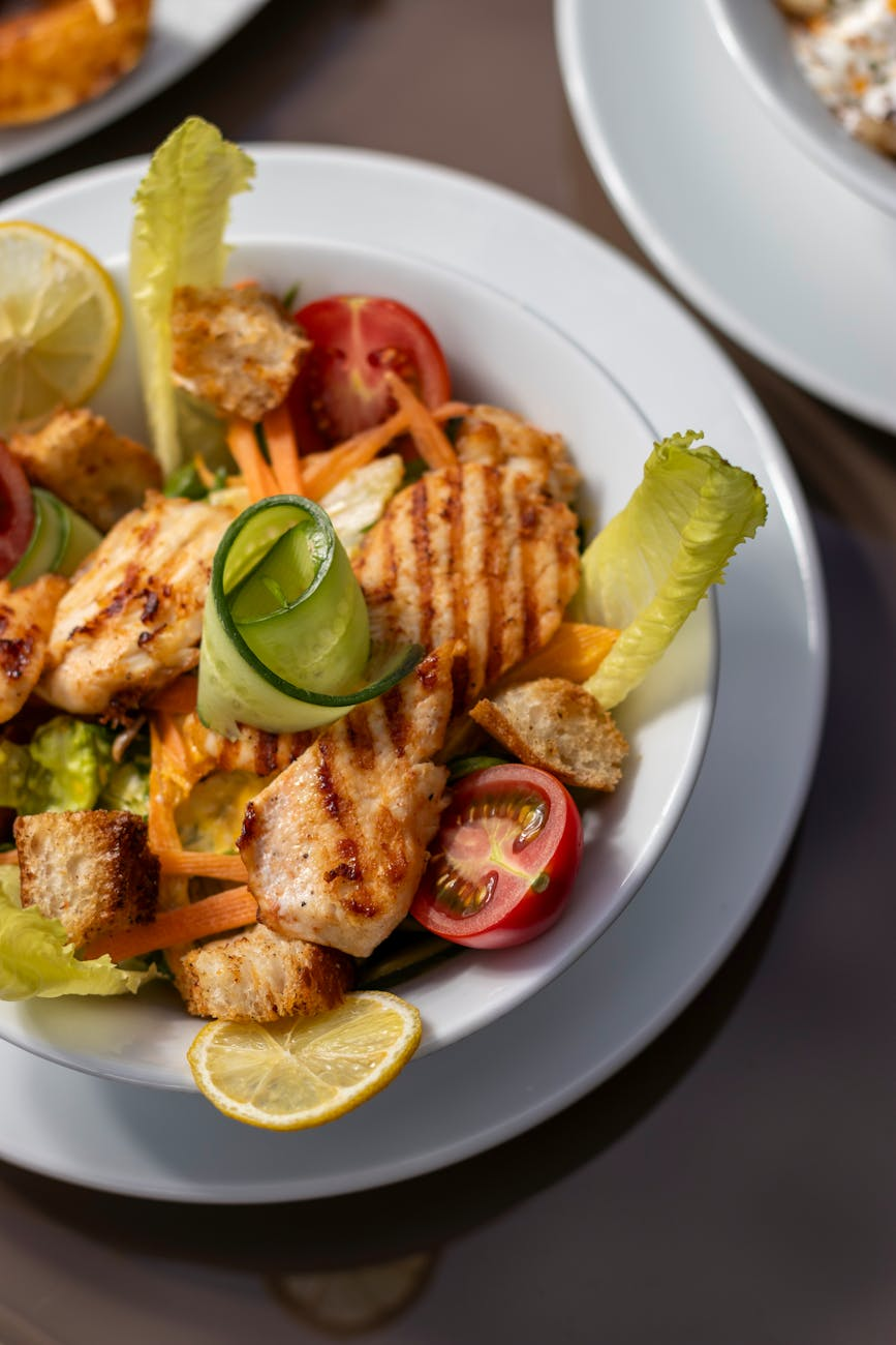 Person reviewing a calorie tracking app on a smartphone next to a kitchen scale, a balanced plate of grilled chicken and roasted vegetables, and a glass of water on a sunlit table